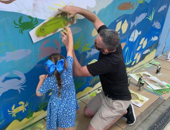 A man helping a little girl spray paint a fish on the wall.